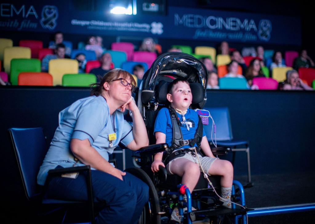 Child in wheelchair and nurse watching a film in the MediCinema with children and parents in seats behind them.